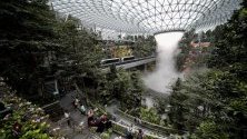 Пътници разглеждат 40-метровия Дъждовния вортекс на летище "Чанги" в Сингапур - най-големият в света водопад в затворено помещение. Построена за над 1 мил. евро, 10-етажната структура свързва терминалите на летището. В нея има хотел, магазини и ресторанти.